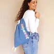 Model smiling wearing Personalised Rainbow Name BagBase Denim Drawstring Bag on back in front of neutral coloured background