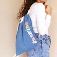 Model holding Personalised Rainbow Name BagBase Denim Drawstring Bag on back with natural coloured background