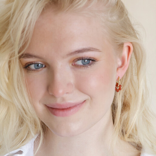 Model looking to side smiling whilst wearing Brick Red Resin Flower Huggie Hoop Earrings in Gold