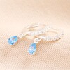 Blue Teardrop Crystal Huggie Hoop Earrings in Silver on top of beige coloured fabric