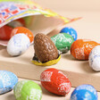 Yellow egg partially unwrapped, surrounded by other flavours in Tony's Chocolonely Egg-stra Special Mix of Chocolate Mini Eggs on Beige Surface