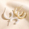 Encased Pearl Huggie Hoop Earrings in Silver with gold version on top of beige coloured fabric