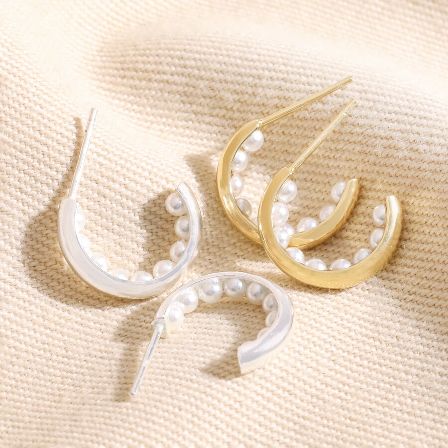 Encased Pearl Huggie Hoop Earrings in gold and silver arranged on top of beige fabric