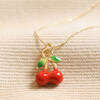 Close Up of Enamel Red Cherry Pendant Necklace in Gold on Beige Fabric