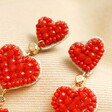 Close up of My Doris Red Beaded Heart Drop Earrings on beige coloured backdrop